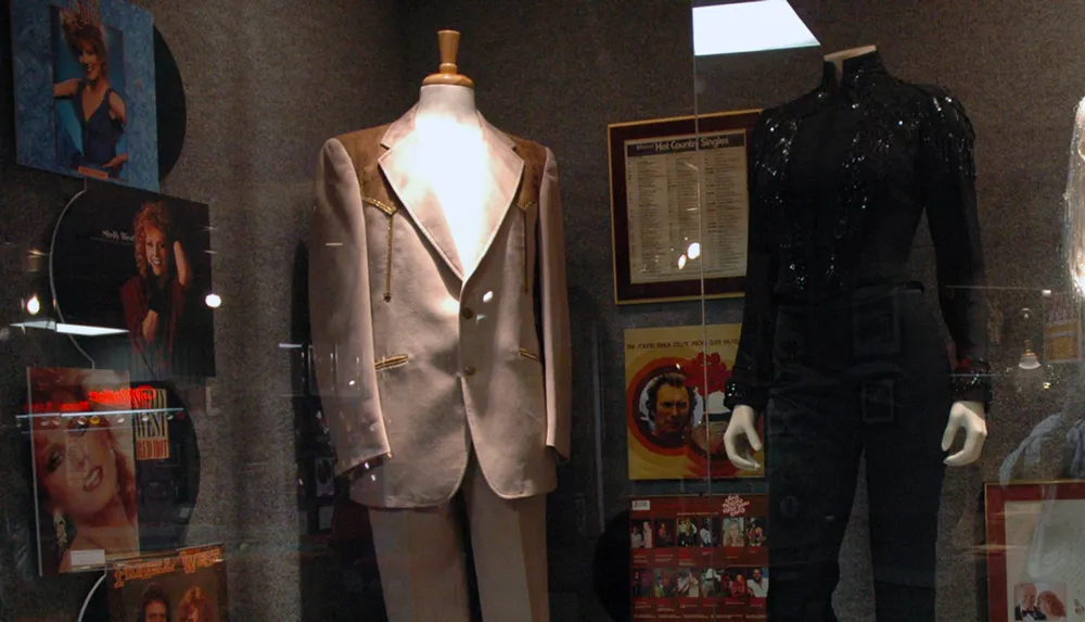 The image shows a display case with two mannequins dressed in a light-colored suit and a black sequined outfit surrounded by album covers and memorabilia likely from a music artist or band