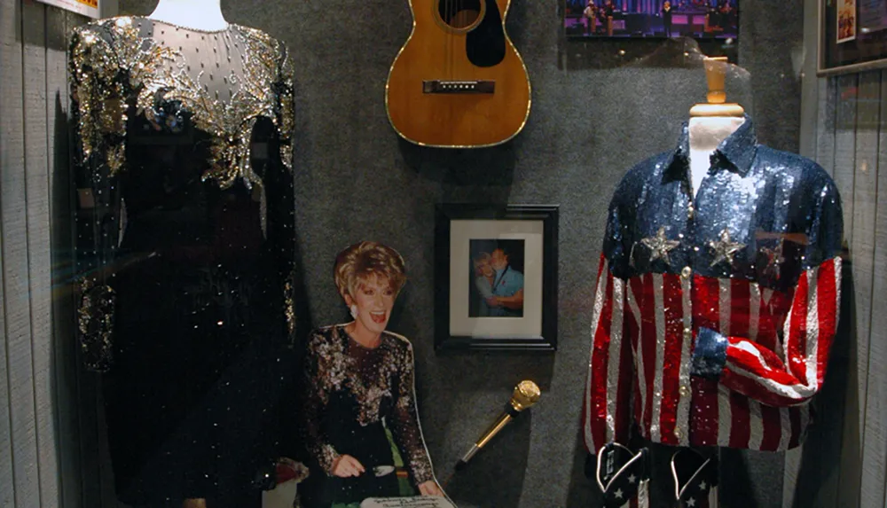 The image showcases a display of sequined performance outfits a guitar framed photographs and a microphone likely part of a music memorabilia collection