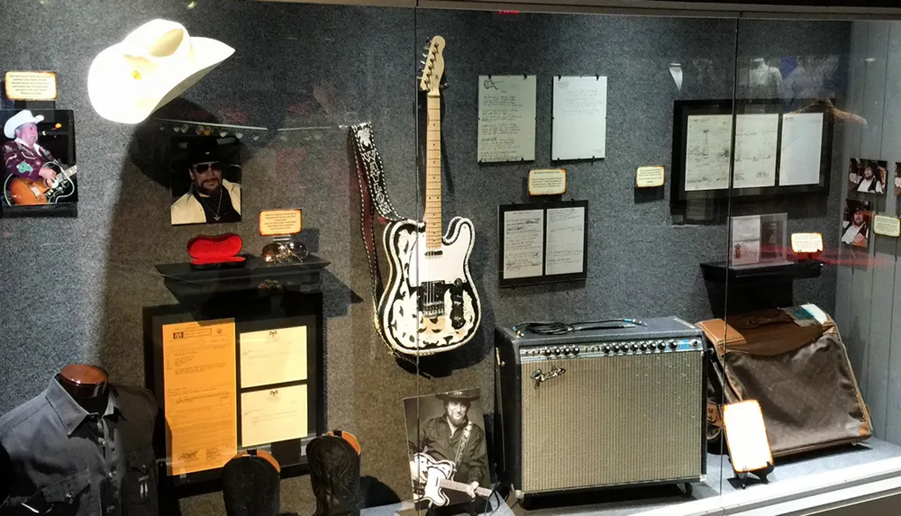 The image shows a display case with various music memorabilia including a decorative guitar photographs clothing a cowboy hat and hand-written notes suggesting a tribute to a musicians career