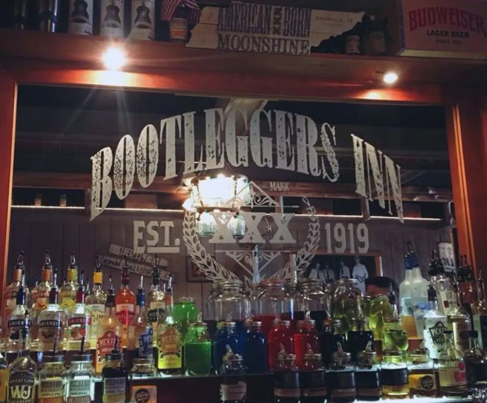 The image shows a bar shelf with an array of colorful liquor bottles in front of a decorative sign that reads Bootleggers Inn Est 1919
