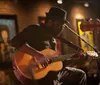 A musician wearing a hat is intently playing an acoustic guitar on a warmly lit stage