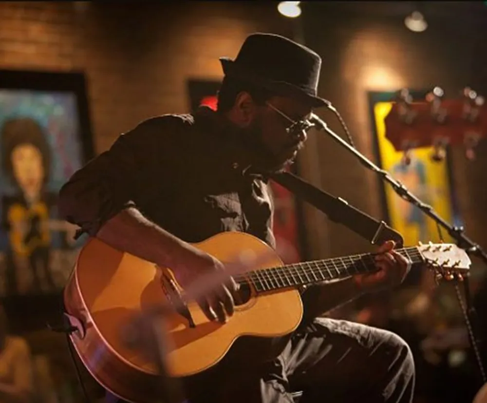 A musician wearing a hat is intently playing an acoustic guitar on a warmly lit stage