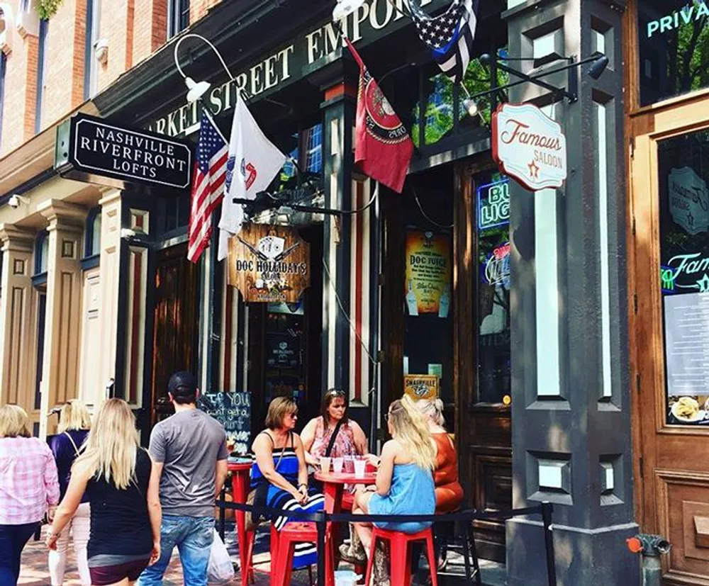 People are walking by and seated outside at a lively city street bar beneath signage that suggests this could be a historic area