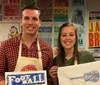 A man and a woman are smiling while holding artwork standing in front of a wall adorned with colorful concert posters