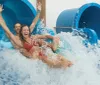 Two people are joyfully sliding down a water chute into a splash pool with their arms raised and expressions filled with excitement