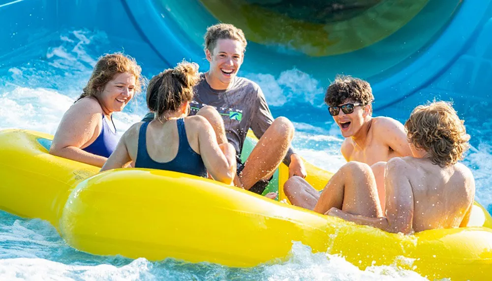 A group of friends is laughing and enjoying a sunlit ride down a water slide in a yellow raft