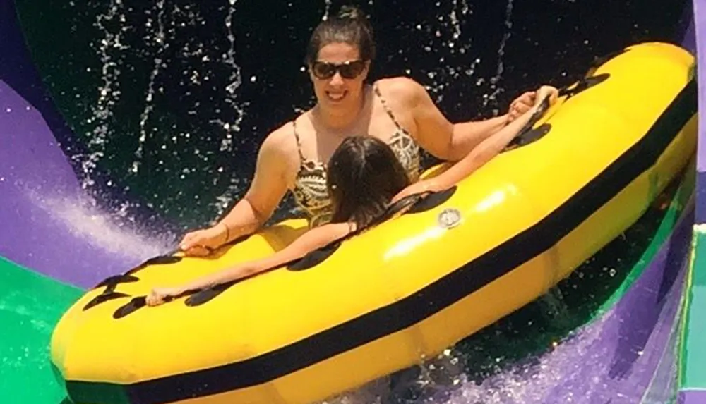 A person wearing sunglasses is sliding down a water slide on a yellow double tube with a child in front
