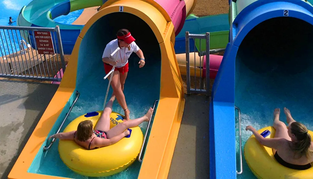 A lifeguard stands by as people launch down colorful water slides on inflatable tubes at an outdoor water park
