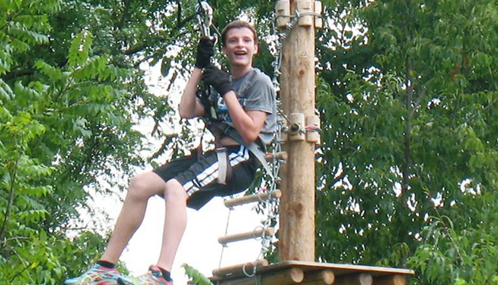 A person wearing safety gear is smiling while sitting on a wooden platform amidst greenery possibly ready for a zip line or rope course activity
