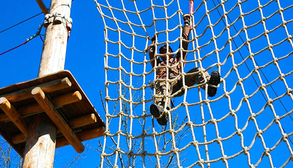 A person is navigating a rope net obstacle at a high ropes adventure course under a clear blue sky