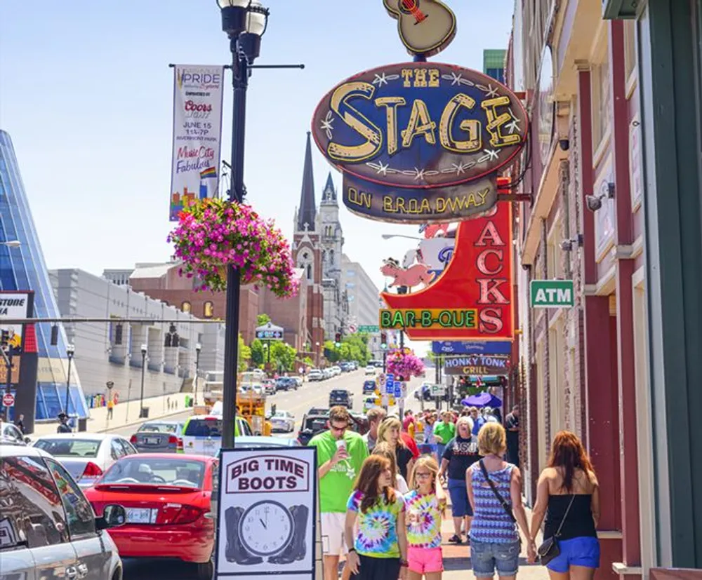 Pedestrians stroll down a vibrant urban street lined with colorful signs and storefronts including one for The Stage on Broadway under a clear blue sky
