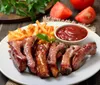 The image shows a plate of barbecued ribs accompanied by french fries and a side of ketchup garnished with a parsley leaf with fresh tomatoes and herbs in the background