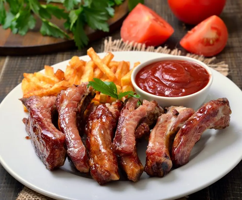 The image shows a plate of barbecued ribs accompanied by french fries and a side of ketchup garnished with a parsley leaf with fresh tomatoes and herbs in the background