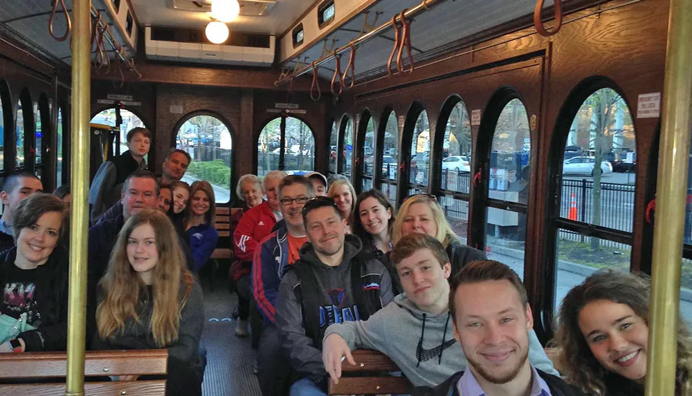 A cheerful group of people is posing for a photo inside a trolley or streetcar with wooden interiors and large windows