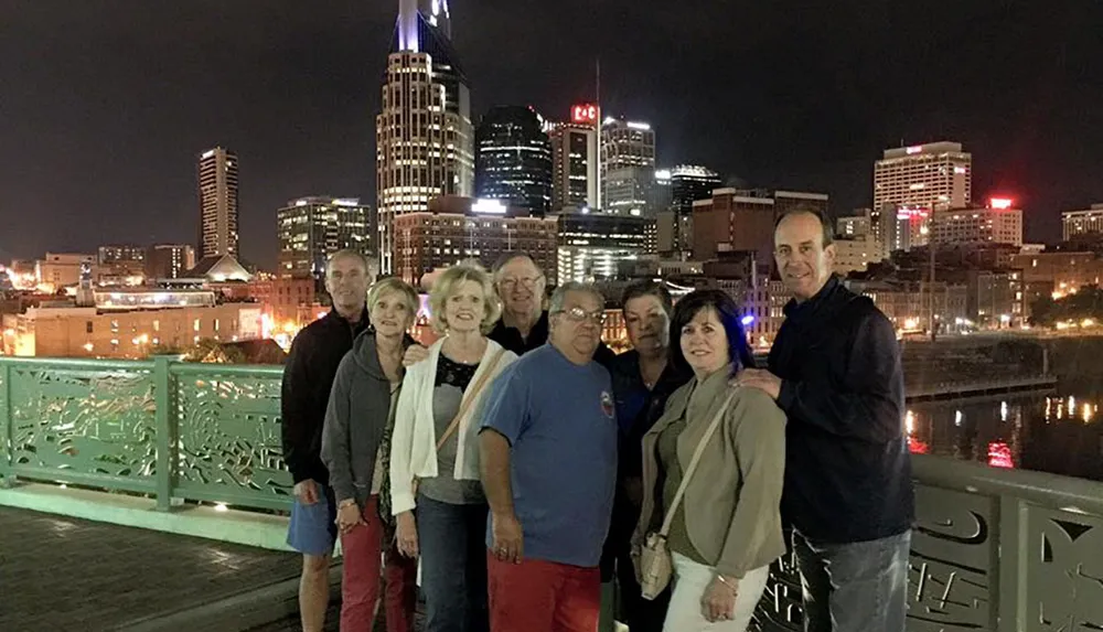 A group of eight people is posing for a photo on a bridge at night with a city skyline illuminated in the background