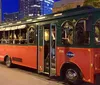 The image shows a red and green trolley-style bus parked on a city street at twilight with buildings and street lights visible in the background