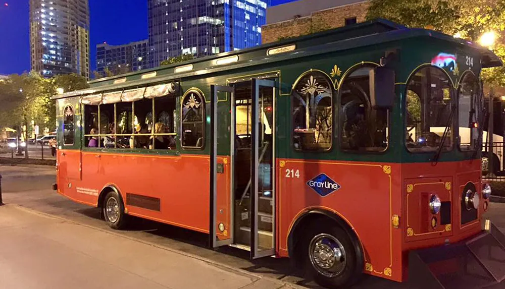 The image shows a red and green trolley-style bus parked on a city street at twilight with buildings and street lights visible in the background