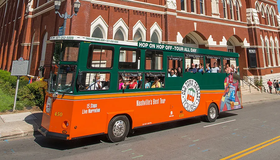 A green and orange trolley bus labeled Old Town Trolley Tours is carrying passengers on a hop-on hop-off tour in a city street.