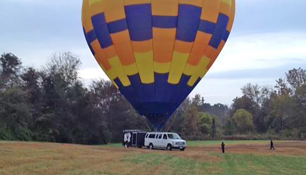 A multicolored hot air balloon is close to touching down in a field while several people are around with a van parked near the balloon