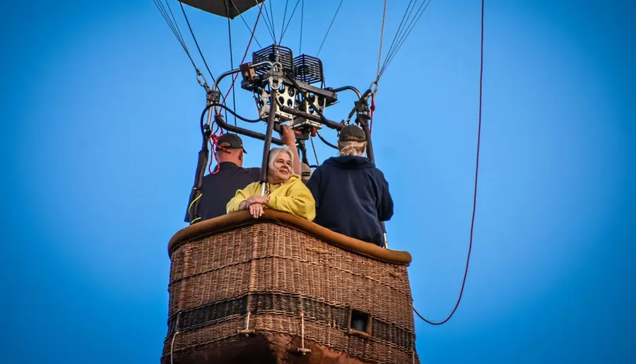 Three people are in a hot air balloon basket against a clear blue sky.