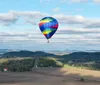 A colorful hot air balloon floats above a scenic landscape with fields trees and rolling hills under a partly cloudy sky