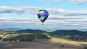 A colorful hot air balloon floats above a scenic landscape with fields, trees, and rolling hills under a partly cloudy sky.