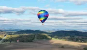 A colorful hot air balloon floats above a scenic landscape with fields, trees, and rolling hills under a partly cloudy sky.