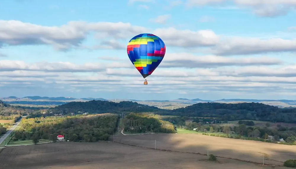 A colorful hot air balloon floats above a scenic landscape with fields trees and rolling hills under a partly cloudy sky