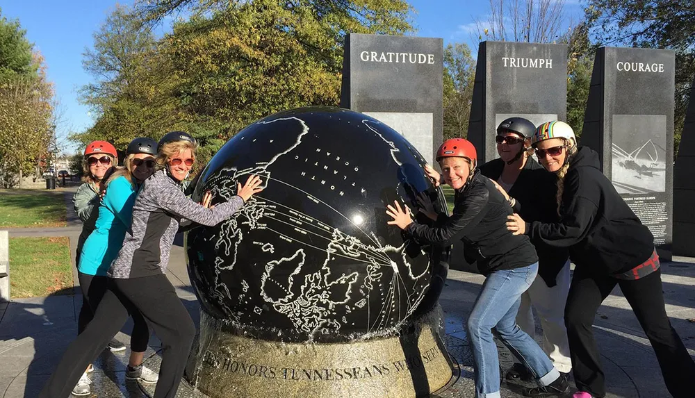 A group of people wearing helmets is playfully posing with a large globe monument outdoors in a park-like setting with plaques bearing words like GRATITUDE TRIUMPH and COURAGE in the background