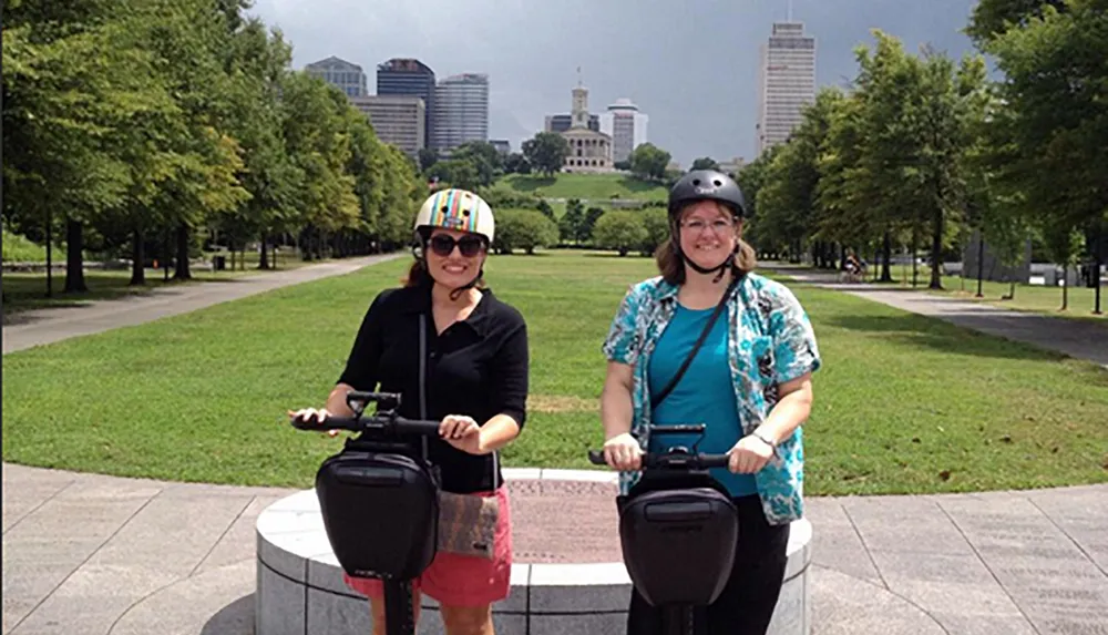 Two people are standing with Segways in a park with trees and city buildings in the background