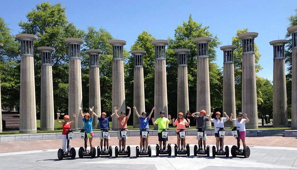 A group of people are posing with their arms raised while standing on Segway personal transporters in front of a row of tall columns