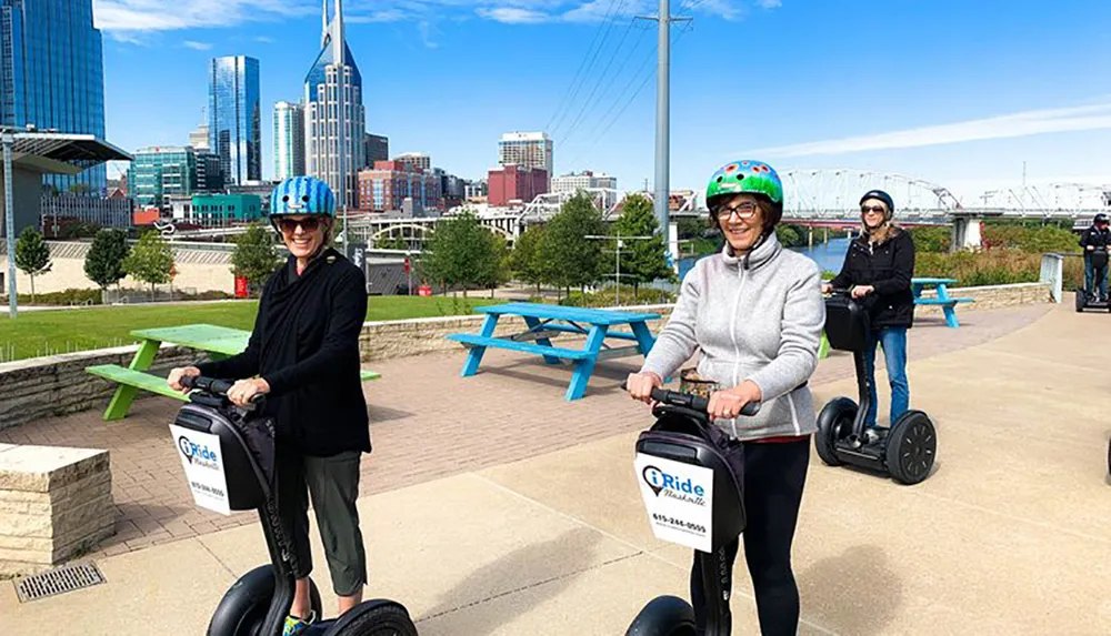 Two individuals are smiling for the camera while riding Segways on a path with the Nashville skyline in the background