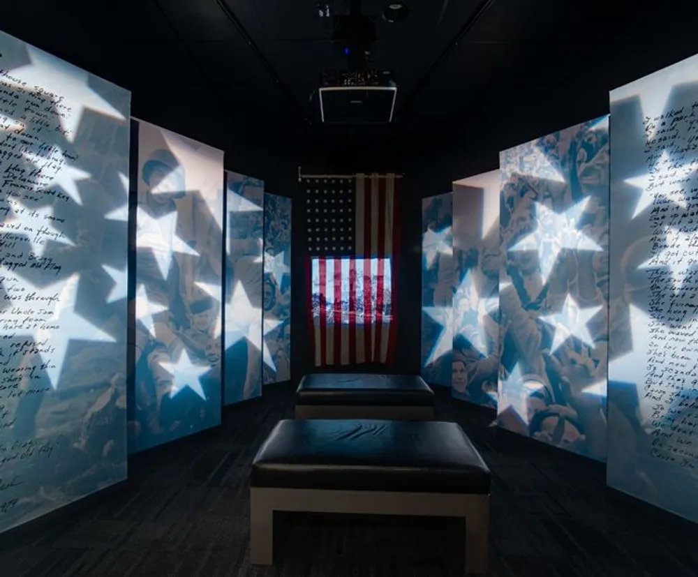 The image depicts a solemn exhibition room with backlit panels featuring handwritten notes and photographs centered around a lit-up American flag creating a contemplative and poignant atmosphere