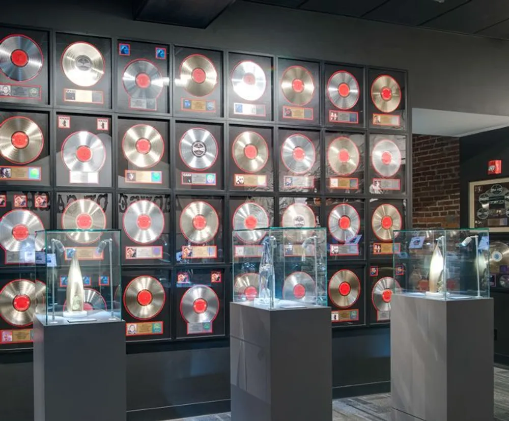 The image shows a wall display of numerous framed gold and platinum record awards with a few award trophies showcased in transparent cases in the foreground