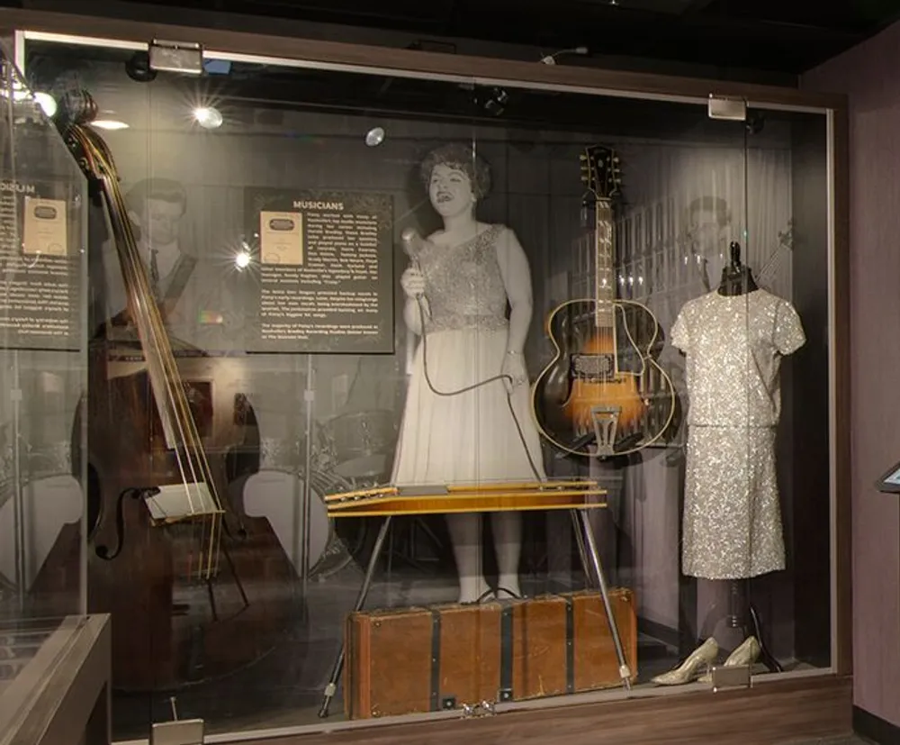 The image shows a museum display featuring a double bass a pedal steel guitar a guitar two mannequins clad in vintage dresses and informational plaques that probably detail the history or significance of the musicians and instruments