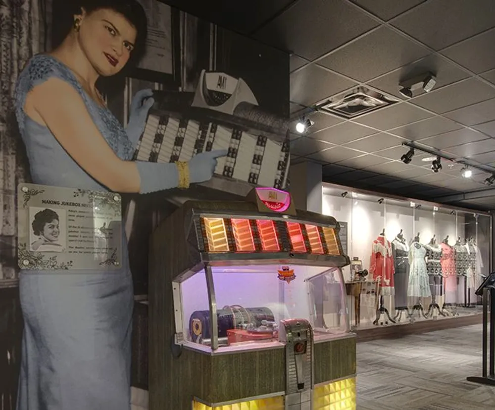 The image shows a vintage-style exhibit with a classic jukebox in the foreground and a life-sized cutout of a woman in a blue dress against a backdrop displaying dresses and memorabilia evoking a nostalgic mid-20th-century vibe