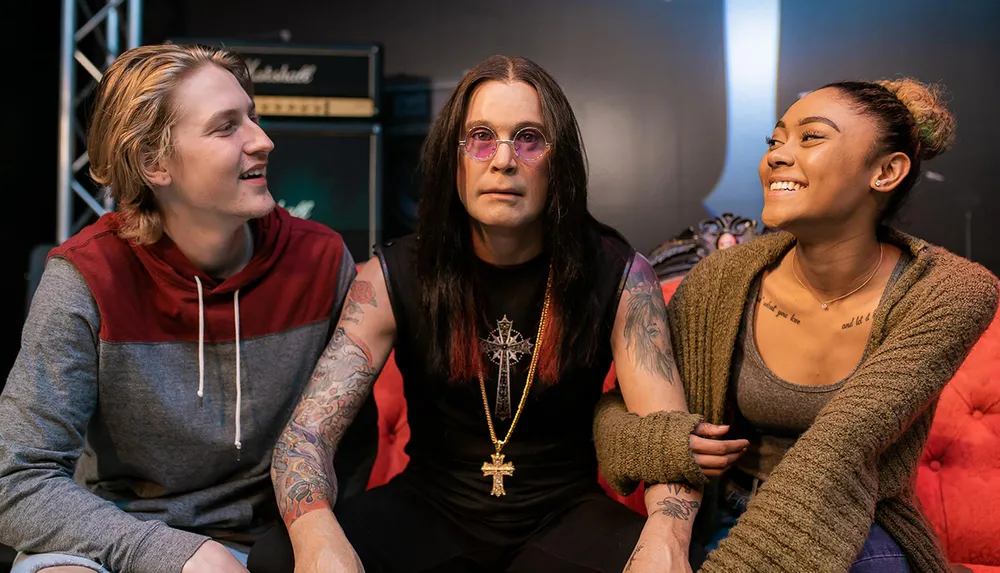 Three people with various tattoos are sitting together and smiling exuding a sense of camaraderie with the central figure wearing circular sunglasses and a crucifix necklace against a backdrop featuring music equipment