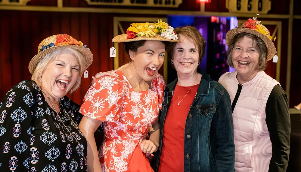 Four cheerful women wearing decorative hats with flowers are posing together with wide smiles conveying a sense of joy and camaraderie