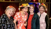Four cheerful women wearing decorative hats with flowers are posing together with wide smiles, conveying a sense of joy and camaraderie.