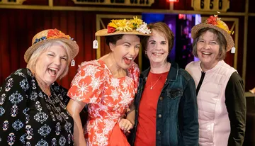 Four cheerful women wearing decorative hats with flowers are posing together with wide smiles, conveying a sense of joy and camaraderie.