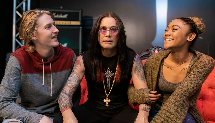 Three people with various tattoos are sitting together and smiling, exuding a sense of camaraderie, with the central figure wearing circular sunglasses and a crucifix necklace, against a backdrop featuring music equipment.
