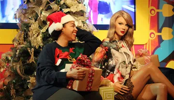 A person wearing a Santa hat and holiday-themed sweater smiles while sitting next to a lifelike wax figure with a Christmas tree and presents in the background.