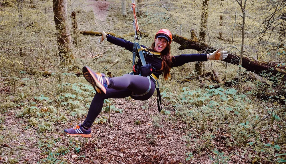 A person is gleefully zip-lining through a forest environment wearing a helmet and safety harness