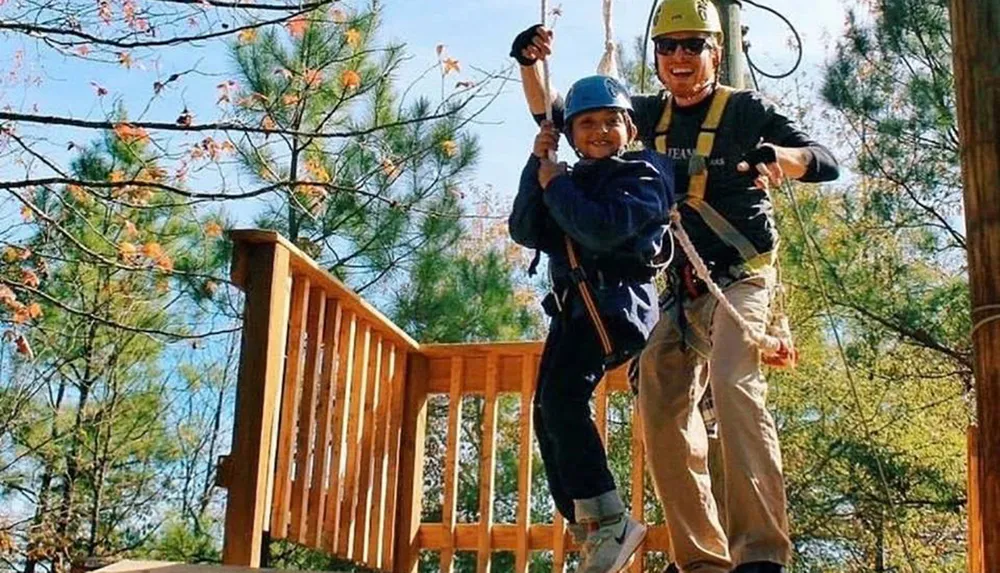 A child and an adult are harnessed and ready to zip line both wearing helmets and cheerful smiles against a backdrop of trees and blue sky