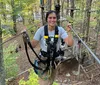 A smiling person is equipped with a harness and safety gear on a high ropes course among trees