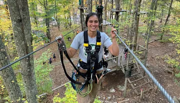 A smiling person is equipped with a harness and safety gear on a high ropes course among trees.