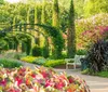 A serene garden pathway is flanked by lush greenery and vibrant flowers with a metal arch and a wooden bench inviting visitors to enjoy the surroundings