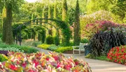A serene garden pathway is flanked by lush greenery and vibrant flowers with a metal arch and a wooden bench inviting visitors to enjoy the surroundings.