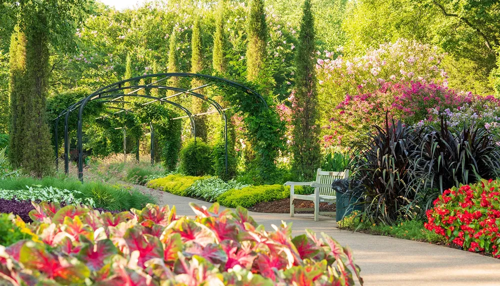 A serene garden pathway is flanked by lush greenery and vibrant flowers with a metal arch and a wooden bench inviting visitors to enjoy the surroundings