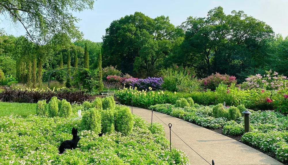 The image shows a beautifully landscaped garden with a variety of colorful flowers and neatly trimmed shrubs alongside a paved walkway with trees in the background under a clear sky